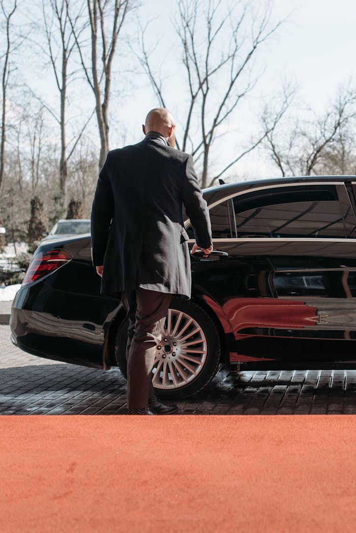 About Bald man in a black suit approaches a luxury black car on a sunny day.